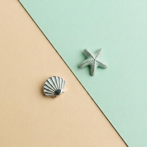 May include: Two enamel pins featuring a sea-themed design. One pin is a light mint green starfish with silver-toned detailing along its arms. The second pin is a fan-shaped seashell with light mint green enamel and silver-toned metal ridges.