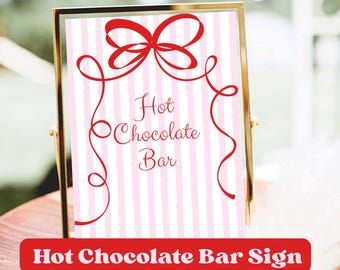 Può includere: Un cartello incorniciato con un bordo dorato mostra il testo "Hot Chocolate Bar" in rosso, adornato con un fiocco rosso e linee decorative. Lo sfondo presenta strisce verticali rosa e bianche. La parte inferiore del cartello recita "Hot Chocolate Bar Sign".