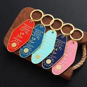 Op de afbeelding: Vijf kleurrijke sleutelhangers met gouden ringen. Elke sleutelhanger heeft een andere kleur: rood, blauw, lichtblauw, donkerblauw en roze. De sleutelhangers hebben de tekst "ADVENTURE AWAITS" en "HOTEL LA More BISBEE ARIZONA".