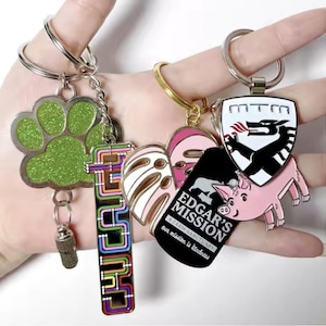 Puede incluir: Una colección de llaveros con varios diseños. Incluye una huella de pata verde brillante, un diseño "CHUCK" colorido, un cerdo rosa y un escudo negro con un dragón. Algunos llaveros tienen texto, incluyendo "EDGAR'S MISSION".