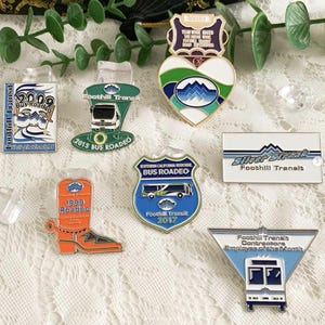 Puede incluir: Una colección de coloridos pines de esmalte con varios diseños y texto. Los pines incluyen un pin en forma de corazón con un diseño de montaña, un pin rectangular con el texto "Silver Streak" y pines con temas relacionados con autobuses.