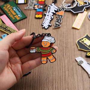 May include: A gingerbread man keyring with a white chef's hat and a mug. The gingerbread man is wearing a red and green sweater. Other keyrings are in the background.