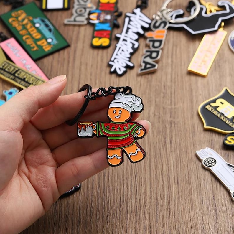 May include: A gingerbread man keyring with a white chef's hat and a mug. The gingerbread man is wearing a red and green sweater. Other keyrings are in the background.