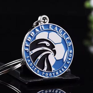 May include: A silver keychain with a circular emblem. The emblem has a blue border and features the text "JEDDAH LADIES FOOTBALL CLUB EAGLES". The design includes a white and black eagle and a football.