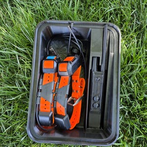May include: A black plastic case containing two orange dog collars with black accents and a black handheld device with an antenna. The collars have silver buckles and are in a black case with a top compartment.