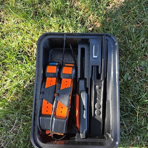 May include: A black storage case holding two orange and black dog training collars, a handheld device, and other accessories. The collars have black straps and orange details. The case is open, showing the contents arranged neatly.