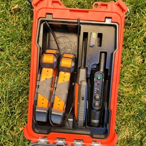 May include: A red waterproof case with a black interior, holding two orange and black dog collars, a black handheld device, and a battery. The case is open on green grass.