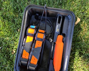 May include: A black plastic tray containing two orange and black tracking collars with antennas and a handheld orange and black GPS device. The items are arranged in molded compartments alongside a black charging cable with a connector.