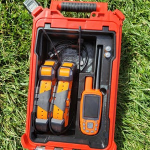 May include: A red case, open to reveal an orange Garmin GPS device, two orange and black collars, and other accessories. The case has a black handle and is on green grass. The Garmin device has a black screen and several buttons.