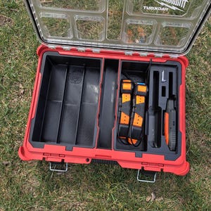 May include: Open red tool case with a clear lid, revealing black compartments and orange and black tools. The case is on a grassy surface. The Milwaukee logo is visible on the lid.