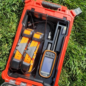 May include: A red waterproof case containing a Garmin Alpha 100 handheld GPS device, two orange and black dog collars, and a black charging cable. The case has a black handle and a clear plastic latch.