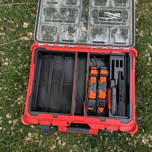 May include: Open red and black tool case with a clear lid, containing various compartments and tools. The case includes orange and black collars, and black electronic devices. The brand name "Milwaukee" is visible on the lid.