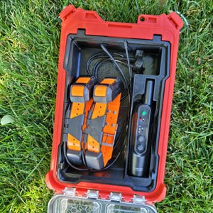 May include: A red, rectangular case with a black interior, holding two orange and black dog training collars, a black handheld device, and charging cables. The case is open, set against a background of green grass.