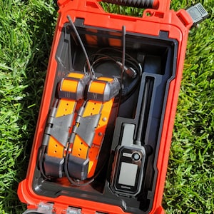 May include: Open red case containing two orange and black dog tracking collars, a handheld GPS device, and related cables. The case is open, revealing the contents against a backdrop of green grass.
