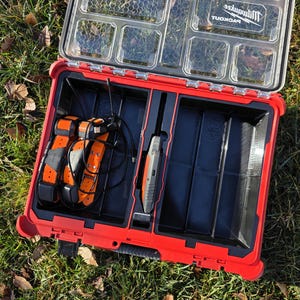 May include: A red and black Milwaukee Packout tool box, open to show compartments and tools. Inside are orange and black collars, a handheld device, and other accessories. The clear lid allows visibility of the contents.