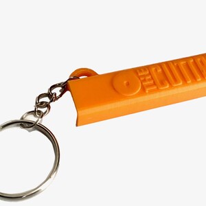 May include: A bright orange plastic keychain featuring a rectangular body with the embossed text THE CUTTR and a circular icon. The orange component is attached to a standard silver-colored metal split ring by a short silver-colored metal chain.