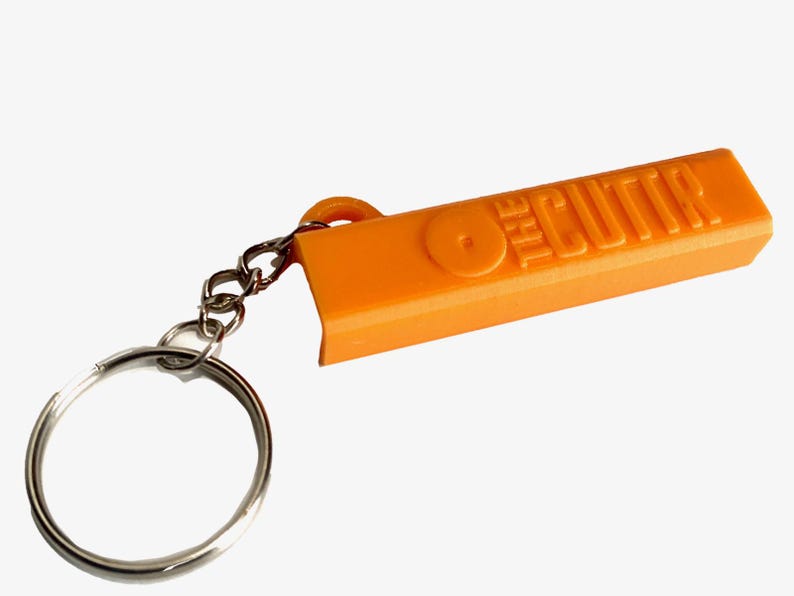 May include: A bright orange plastic keychain featuring a rectangular body with the embossed text THE CUTTR and a circular icon. The orange component is attached to a standard silver-colored metal split ring by a short silver-colored metal chain.