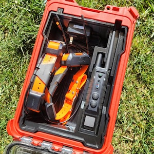 May include: A red hard case with a black and orange dog collar, a black and orange dog training collar, a black handheld device with a screen and buttons, and a black charger.