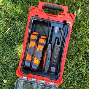 May include: A red, rectangular carrying case with a black interior, containing two orange and black dog collars, a handheld device, and other accessories. The case is open, revealing the contents, and sits on green grass.