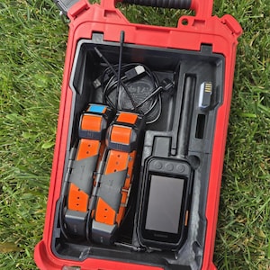 May include: A red hard-shell carrying case containing two orange and black Garmin tracking collars, a black handheld GPS device with an antenna, a black charging cable, and a small rectangular component, all organized within a custom-molded interior.
