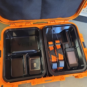May include: An open orange YETI case with black interior compartments. The left side contains a battery and a device with a QR code. The right side holds two orange and black dog tracking collars and a handheld GPS device.