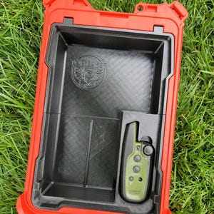 May include: Open red and black storage case with a green Garmin device inside. The case has a black interior with the words "TOP LAYER" and a logo. The device has buttons and a dial.