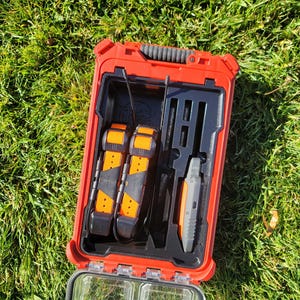 May include: Open red case with black interior, containing two orange and black devices, and a grey device. The case is open, revealing the contents. The case is on a bed of green grass.