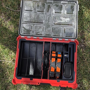May include: Open red and black tool organiser with a clear lid. The organiser contains compartments, straps, and a black device. The lid has the brand name "Milwaukee" printed on it.