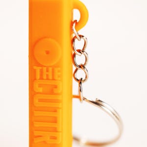 May include: A bright orange rectangular keychain featuring the embossed text THE CUTTR on its front surface. The object includes a silver-colored metal chain and a circular split ring attached to a small loop at the top right side of the orange body.