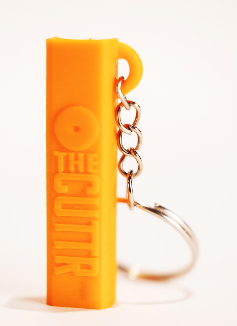 May include: A bright orange rectangular keychain featuring the embossed text THE CUTTR on its front surface. The object includes a silver-colored metal chain and a circular split ring attached to a small loop at the top right side of the orange body.