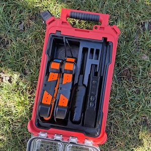 May include: A red, rectangular carrying case with a black handle and a clear, honeycomb-patterned compartment. Inside, there are two orange and black devices, along with two black devices. The case is open, revealing the contents.