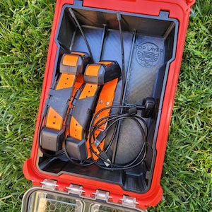 May include: A red, rectangular storage case with a clear, divided compartment and a black interior. Inside, two orange and black electronic collars and a charging cable are visible. The case is open, revealing the contents, and sits on green grass.