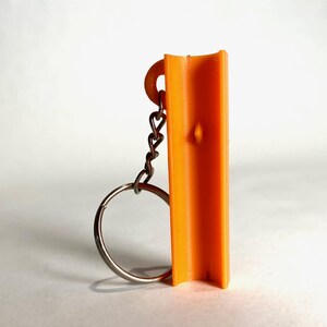 May include: A bright orange, vertically oriented, U-shaped plastic component attached to a silver-colored metal keychain ring. The object features a small, centered protrusion on its inner surface and a circular loop at the top for the chain.