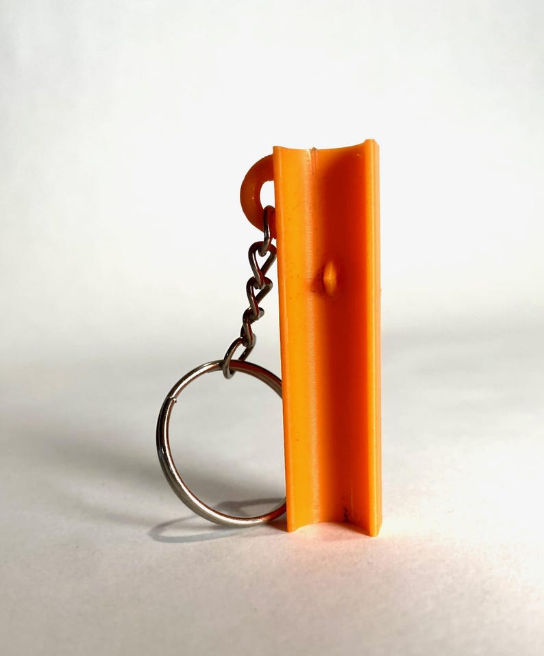 May include: A bright orange, vertically oriented, U-shaped plastic component attached to a silver-colored metal keychain ring. The object features a small, centered protrusion on its inner surface and a circular loop at the top for the chain.