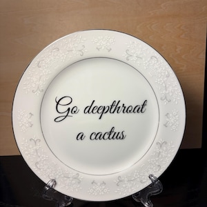 May include: A white ceramic plate with a silver-toned floral pattern along the rim. The center of the plate features the black text "Go deepthroat a cactus" written in a cursive script font. The plate is displayed on a clear plastic stand.