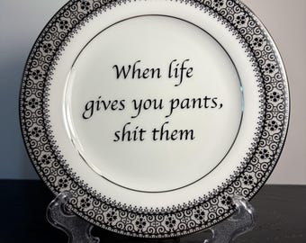 May include: A white ceramic plate with a black decorative border featuring an intricate geometric pattern. The center of the plate displays the black text: When life gives you pants, shit them. The plate is held by a clear plastic stand.