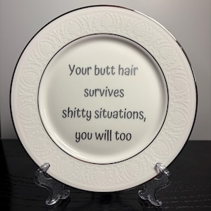 May include: A white ceramic plate with a silver rim and an embossed leaf pattern along the border. The center of the plate features black text that reads: Your butt hair survives shitty situations, you will too. The plate is round and smooth.