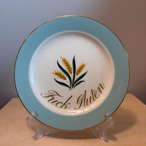 May include: A round white ceramic plate with a light blue border and a gold rim. The center features a graphic of yellow wheat stalks with green leaves. Below the wheat, the text "Fuck Gluten" is written in a gold cursive script font.