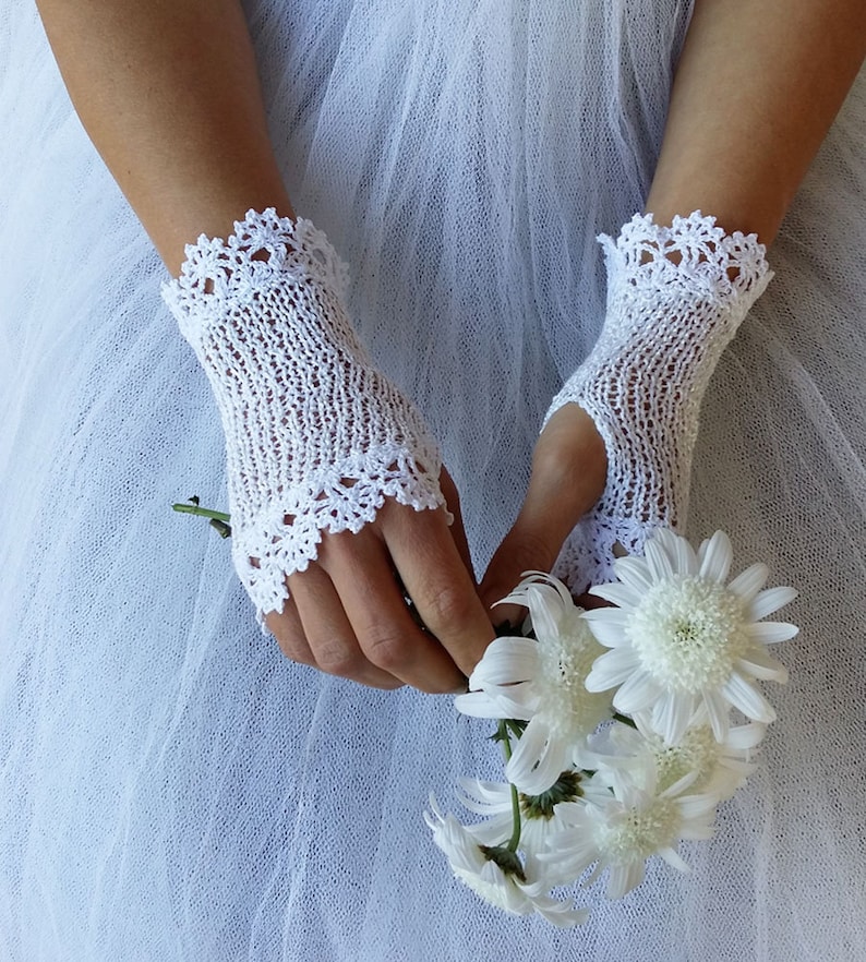May include: A pair of white fingerless gloves crafted from delicate knit fabric, featuring intricate scalloped lace trim at the wrist and knuckle openings. The gloves are worn while holding a small bouquet of white daisy-like flowers.