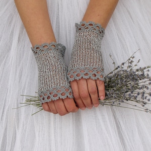 May include: A pair of silver-gray fingerless gloves with a delicate, scalloped crochet trim. The gloves are made of a knitted material and are shown against a white background. The gloves are holding a small bouquet of lavender.