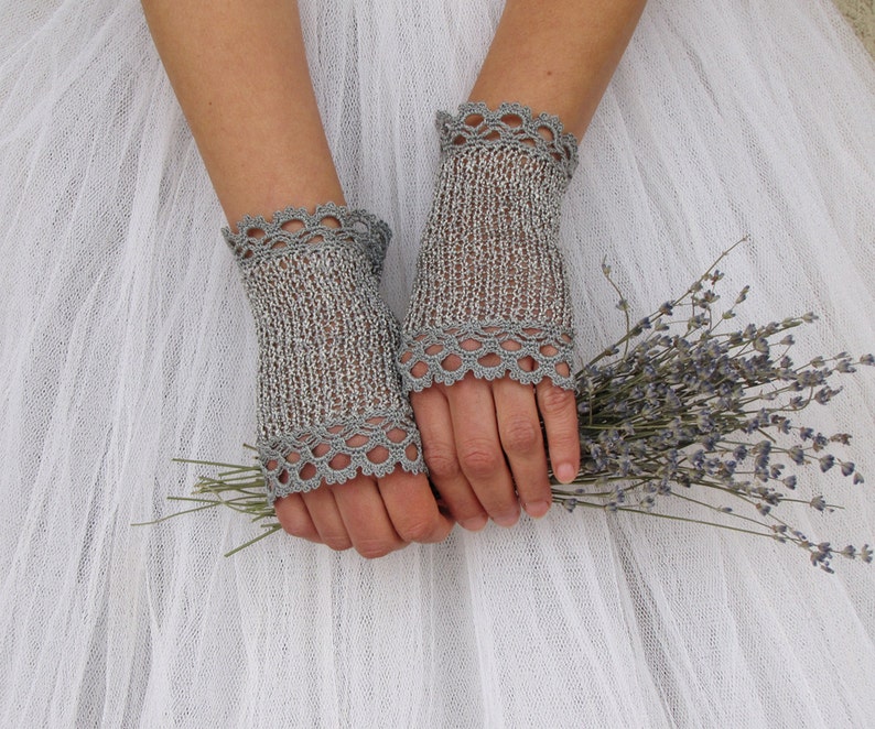 May include: A pair of silver-gray fingerless gloves with a delicate, scalloped crochet trim. The gloves are made of a knitted material and are shown against a white background. The gloves are holding a small bouquet of lavender.
