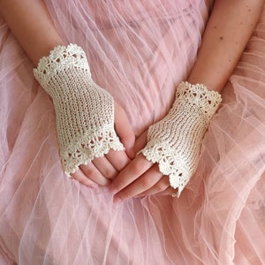 May include: Cream-colored fingerless gloves with a delicate, openwork crochet design. The gloves feature a scalloped edge and are made of a fine, knitted material. The gloves are shown against a soft pink background, highlighting the intricate details of the handmade accessory.