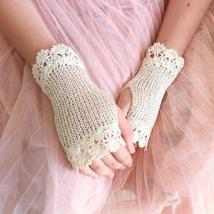 May include: A pair of ivory-colored fingerless gloves with a delicate crochet design. The gloves feature a knitted body and a scalloped crochet trim around the wrist and fingers. The background is a soft pink.