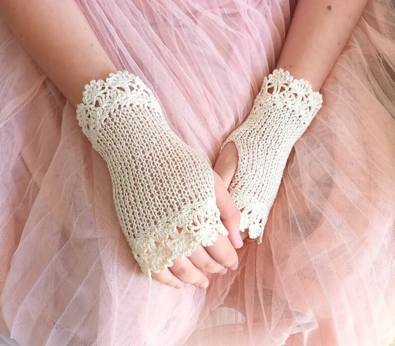 May include: A pair of ivory-colored fingerless gloves with a delicate crochet design. The gloves feature a knitted body and a scalloped crochet trim around the wrist and fingers. The background is a soft pink.
