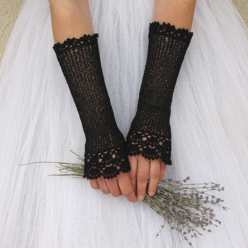 May include: A pair of black fingerless gloves featuring a fine knit texture and intricate scalloped crochet lace detailing at the top and bottom edges. The gloves extend from the knuckles to the mid-forearm, showcasing a delicate openwork pattern.