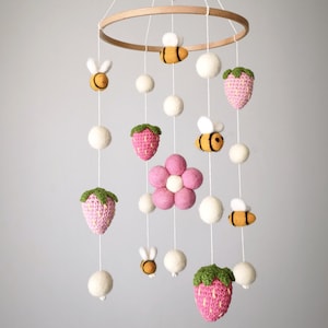 May include: A baby mobile featuring a wooden ring with hanging white felt balls, pink strawberries, yellow and black bees, and a pink flower. The strawberries have green tops. The mobile is suspended against a gray background.