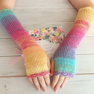 Peut inclure: Une paire de mitaines sans doigts en tricot arc-en-ciel avec un bord festonné. Les mitaines sont faites avec un dégradé de couleurs, commençant par le bleu clair et passant au jaune, au rose et au violet.