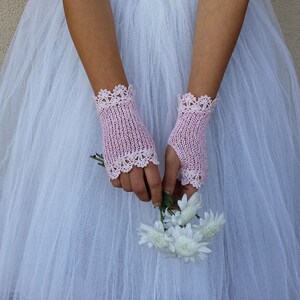 May include: A pair of pale pink fingerless gloves featuring a delicate, intricate crocheted lace trim at both the top and bottom edges. The gloves are crafted with a fine, open-knit texture and are worn over a white tulle skirt while holding flowers.