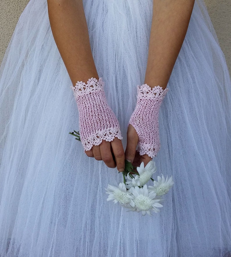 May include: A pair of pale pink fingerless gloves featuring a delicate, intricate crocheted lace trim at both the top and bottom edges. The gloves are crafted with a fine, open-knit texture and are worn over a white tulle skirt while holding flowers.