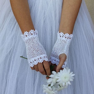 May include: A pair of white fingerless gloves featuring a delicate knit texture and intricate scalloped lace trim at the top and bottom edges. The gloves are crafted from a fine white thread and are worn over a white tulle fabric background.